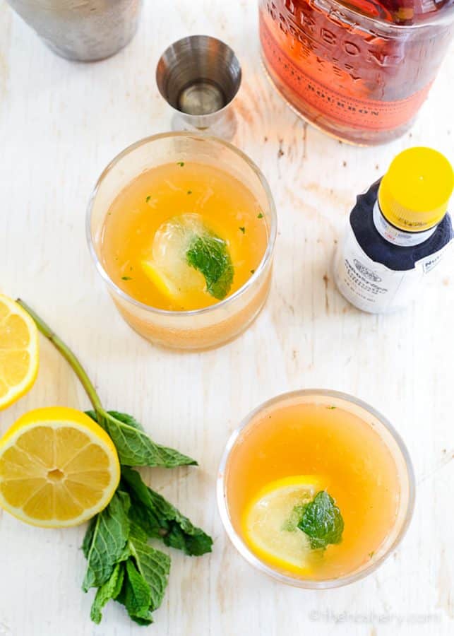 Whiskey Smash - Citrus + Mint Whiskey Cocktail | Overhead view of two glasses of whiskey smash cocktail on white surface. With lemon and mint in the picture. | The Noshery