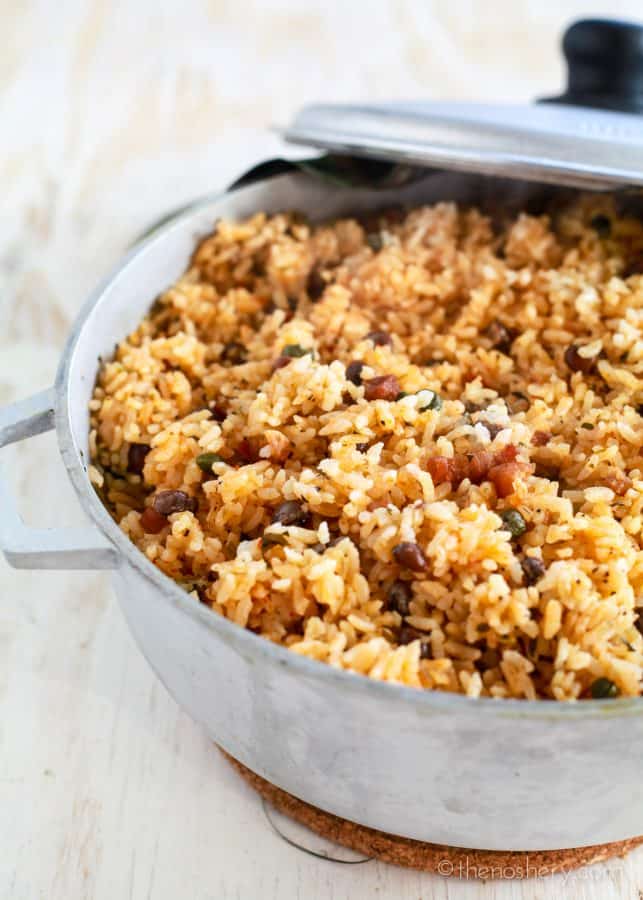 Arroz con Gandules (Puerto Rican Style Rice and Pigeon Peas) | Large pot of orange colored rice with small dark beans and bacon. 