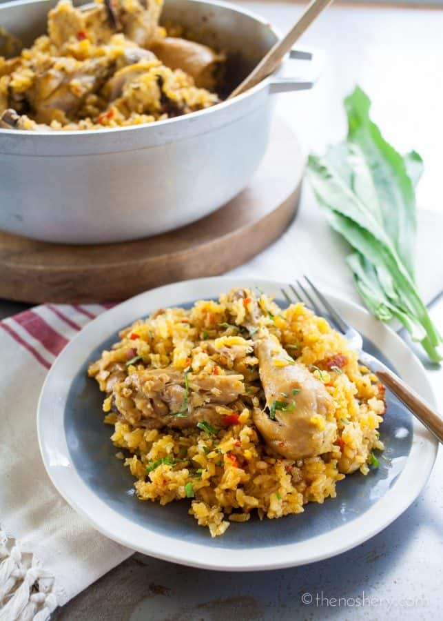 Arroz con Pollo | Side view of chicken and yellow rice on a plate with a large pot in the background. 